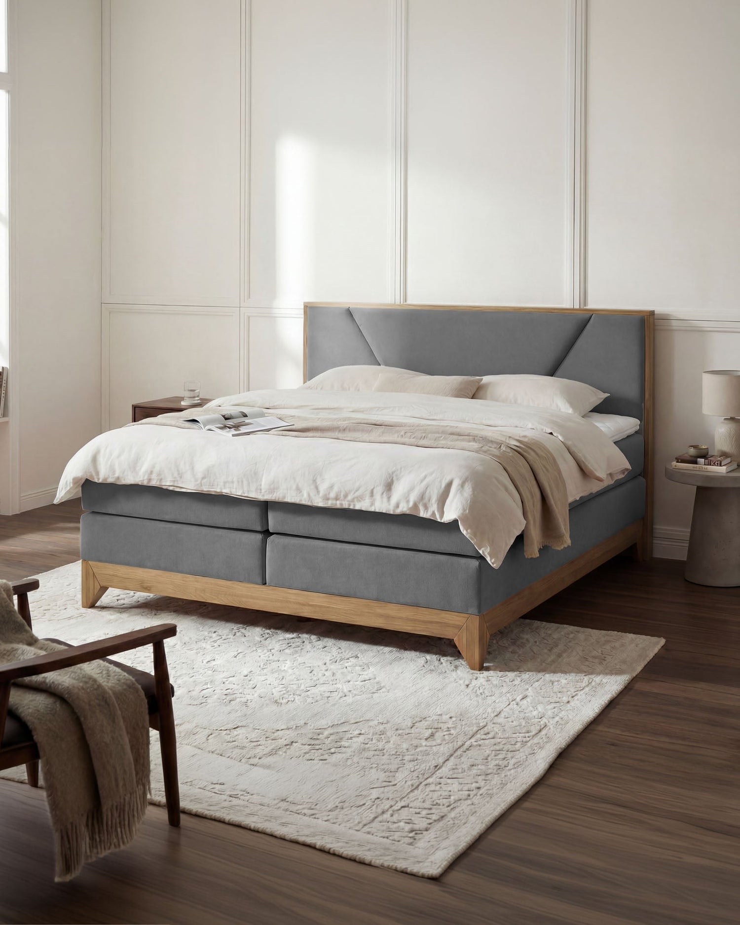 Graues Boxspringbett mit gepolstertem Kopfteil, hellem Holzrahmen und weißer Bettwäsche in einem hellen Schlafzimmer