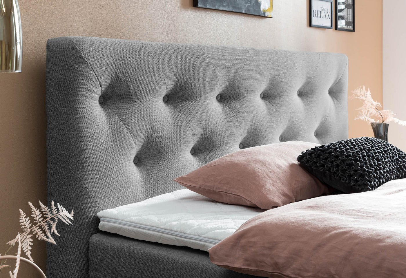 Graues, gestepptes Kopfteil eines Boxspringbettes, weißer Topper und roséfarbene Bettwäsche mit schwarzem Zierkissen
